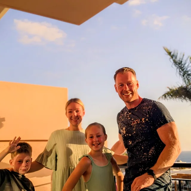 The family on a hotel balcony at golden hour: Matthew perched on the rail with his wife and two kids in sage green, palm tree and sea behind.