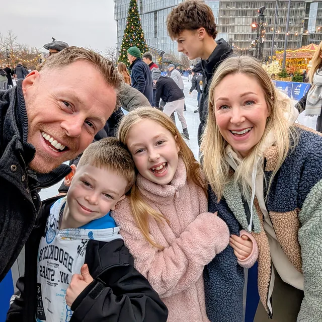 Family selfie at an outdoor ice rink: Matthew grinning with his two kids and wife, a big Christmas tree and carousel behind.