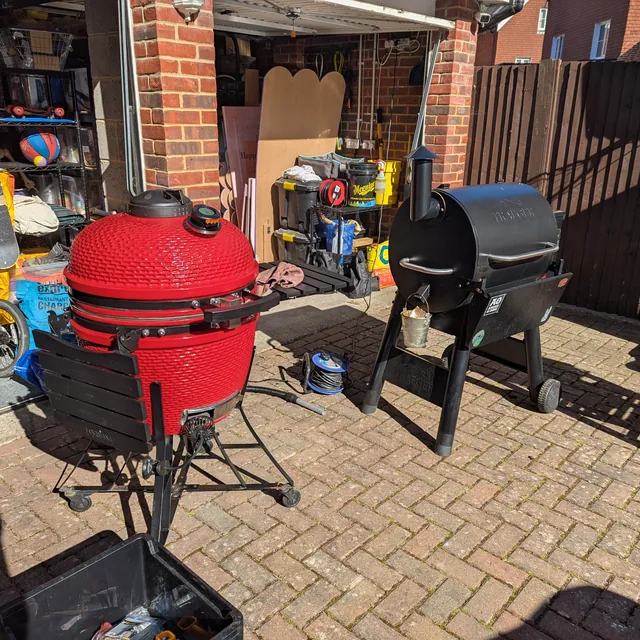 A red ceramic kamado and a black Traeger pellet grill side by side on a brick driveway outside the garage.